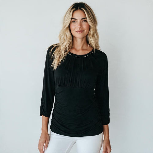 A woman with blonde hair wearing a black three-quarter sleeve top with ruching and white pants, standing against a light gray background.