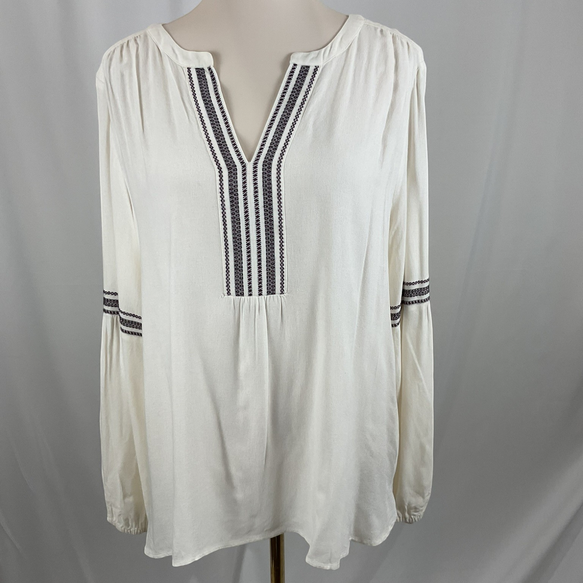 An off-white long-sleeve blouse with black and white striped embroidery on the V-neckline and cuffs, displayed on a mannequin torso against a light gray fabric backdrop.