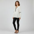 A woman modeling a white long-sleeve blouse with thin black stripes on the chest and cuffs, paired with black skinny pants and black high-heeled sandals, against a light gray studio background.
