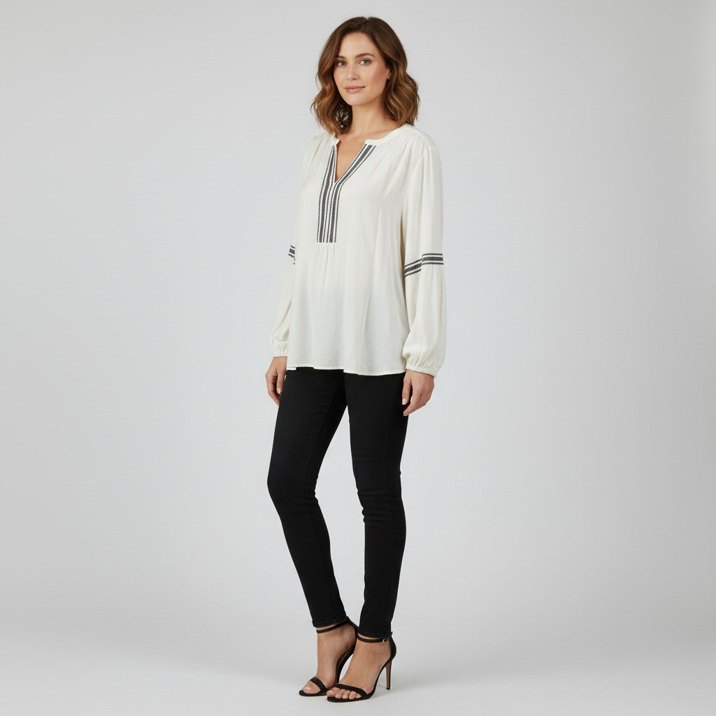 A woman modeling a white long-sleeve blouse with thin black stripes on the chest and cuffs, paired with black skinny pants and black high-heeled sandals, against a light gray studio background.