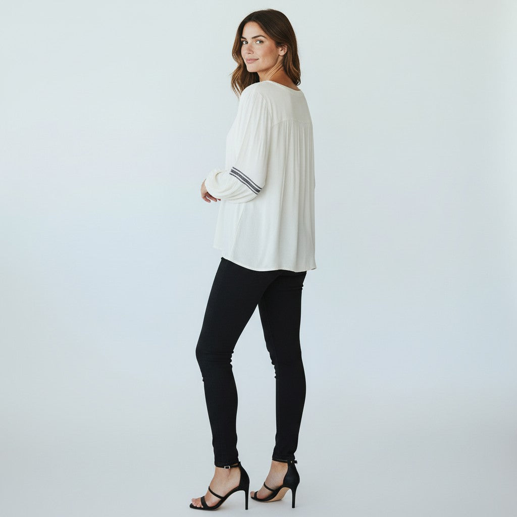 A woman modeling a white long-sleeve blouse with striped cuffs, paired with black skinny pants and black strappy heels, photographed against a light gray studio background.