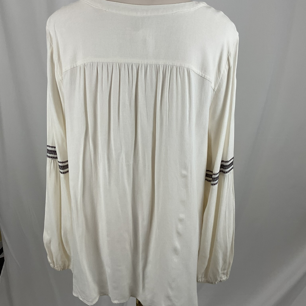 An off-white long-sleeve blouse with a gathered yoke and striped detailing on the sleeves, shown on a mannequin against a light gray backdrop.