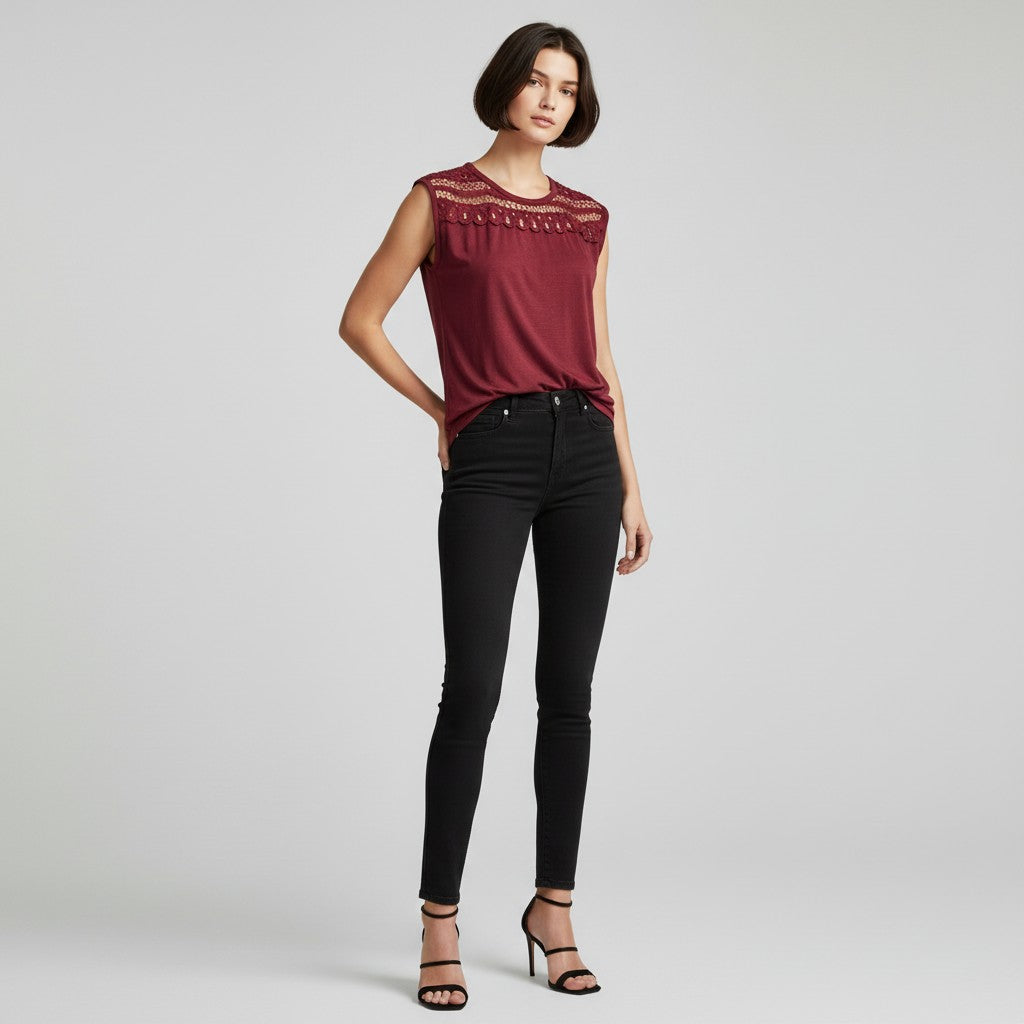 A model wearing a sleeveless burgundy top with a lace yoke and black skinny jeans, standing against a light gray studio background.